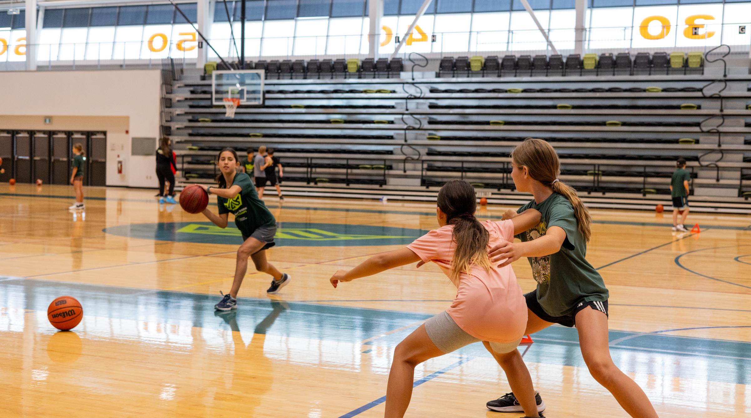 An action shot of a girl in a gymnasium passing a basketball to a teammate who is being guarded by another girl