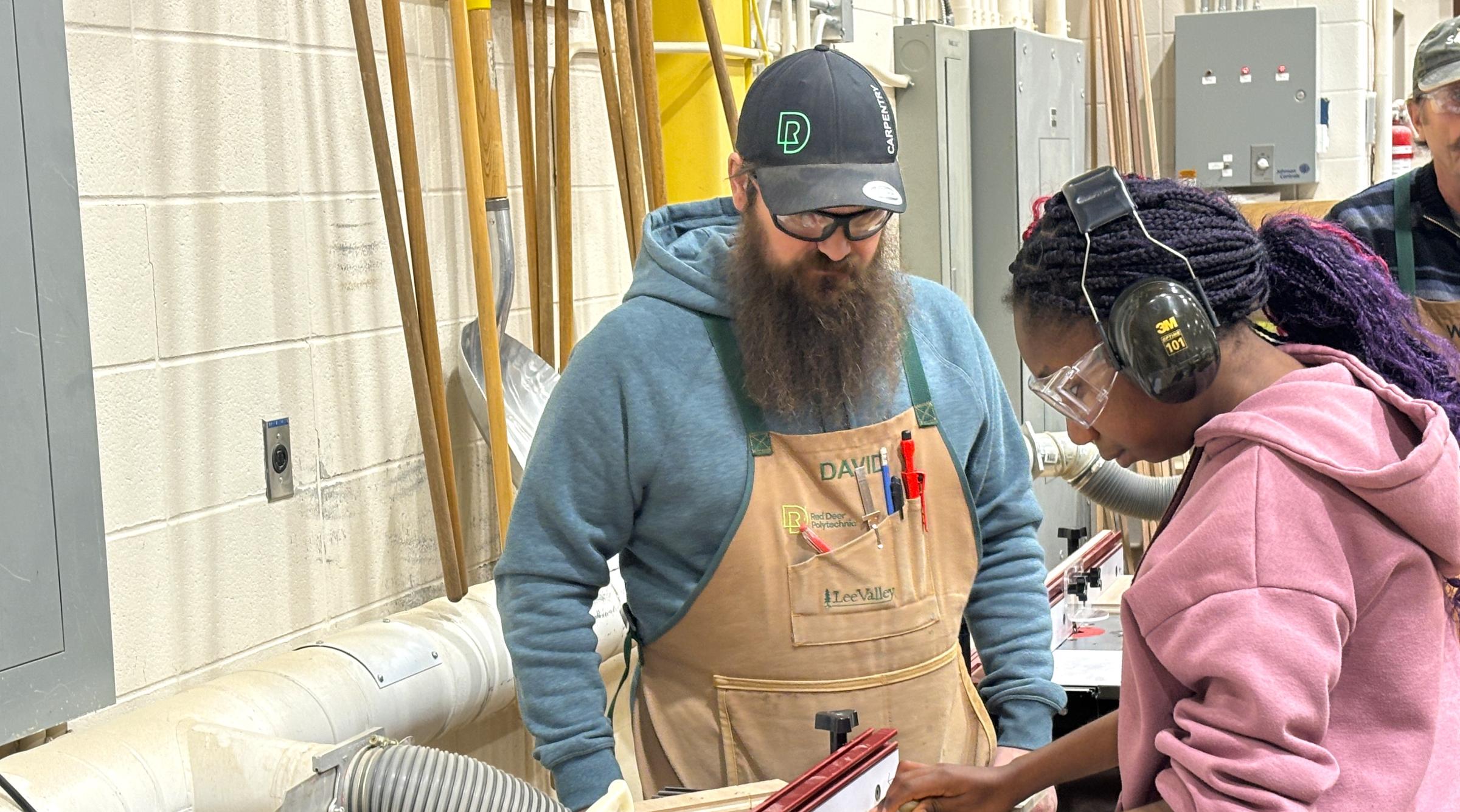 A bearded faculty member watches as a young female student cuts some wood on a large table saw.
