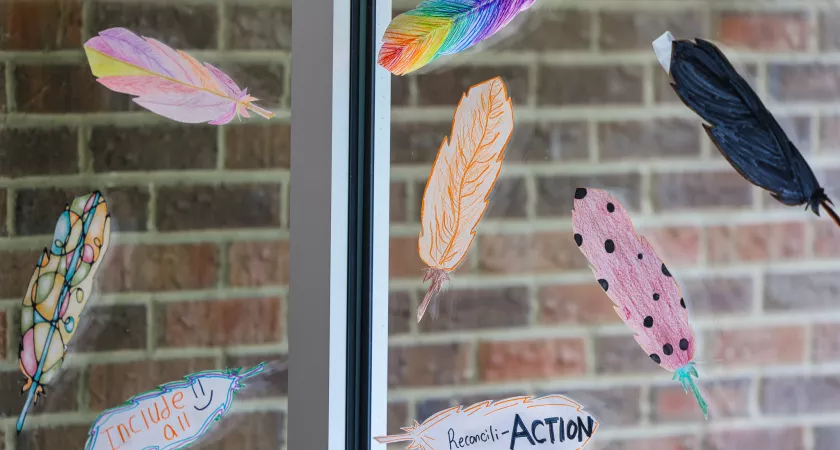 Paper feathers colored with designs and words on a window on campus. The words read "include all" and "reconcili-action"